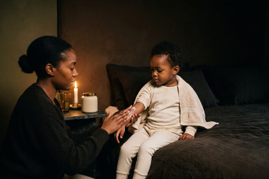 A mother kneeling beside her young child after a bath, gently smoothing moisturiser into the child's forearm by candlelight